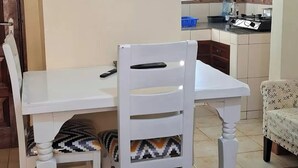 A dining area with a white table and chairs, a kitchen in the background, and a patterned rug on the floor.