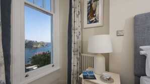 A room with a view of a coastal landscape, a bedside table with a lamp and a book, and a framed artwork on the wall. - Dolly's Plaice -  a cottage that sleeps 4 guests  in 2 bedrooms (Fowey)