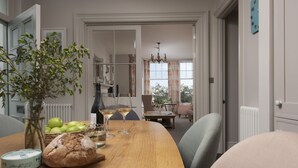 A dining area with a wooden table, two chairs, a vase with green apples, a loaf of bread, and two glasses of white wine. - Dolly's Plaice -  a cottage that sleeps 4 guests  in 2 bedrooms (Fowey)