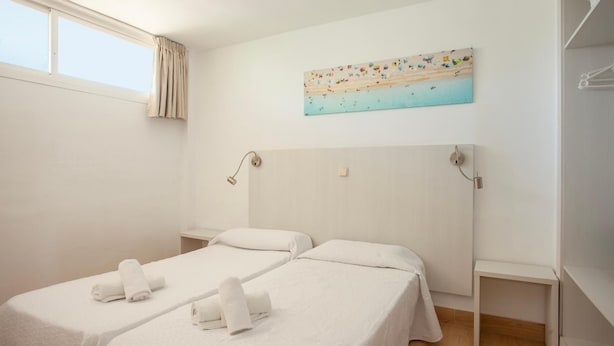 A hotel room with two beds, a painting of a beach scene, a window with curtains, and a wooden floor. - Fiesta Beach Apartments (Sant Llorenc des Cardassar)