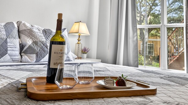 Une table de chevet en bois sur laquelle se trouvent une bouteille de vin Biltmore, deux verres et une assiette avec de la nourriture.