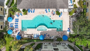 Vue aérienne d’une piscine sur le toit, entourée de nombreuses chaises longues et de parasols, ainsi que de verdure et d’immeubles adjacents.