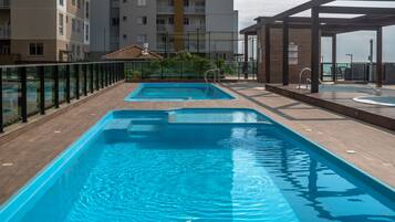 Uma piscina no terraço com deck de madeira, rodeada por guarda-vidro e um edifício ao fundo.