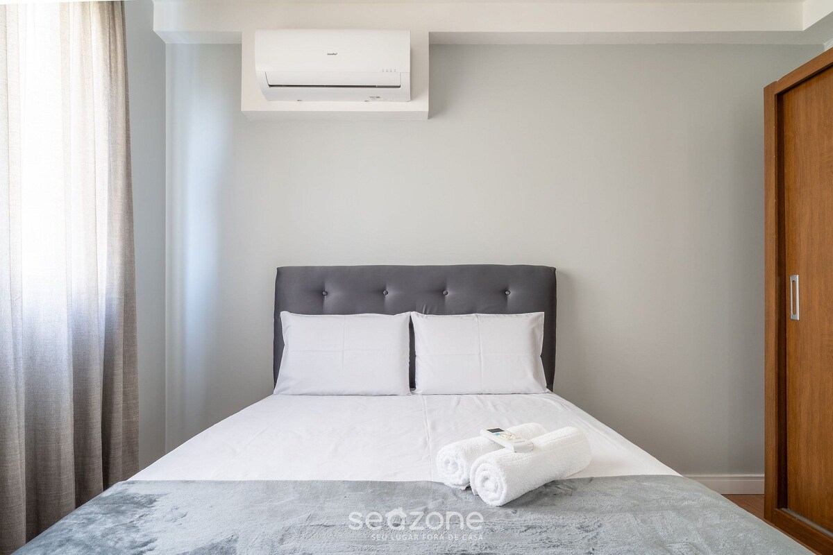 A neatly made bed with white linens and a grey blanket, a grey upholstered headboard, and a white air conditioning unit mounted on the wall.