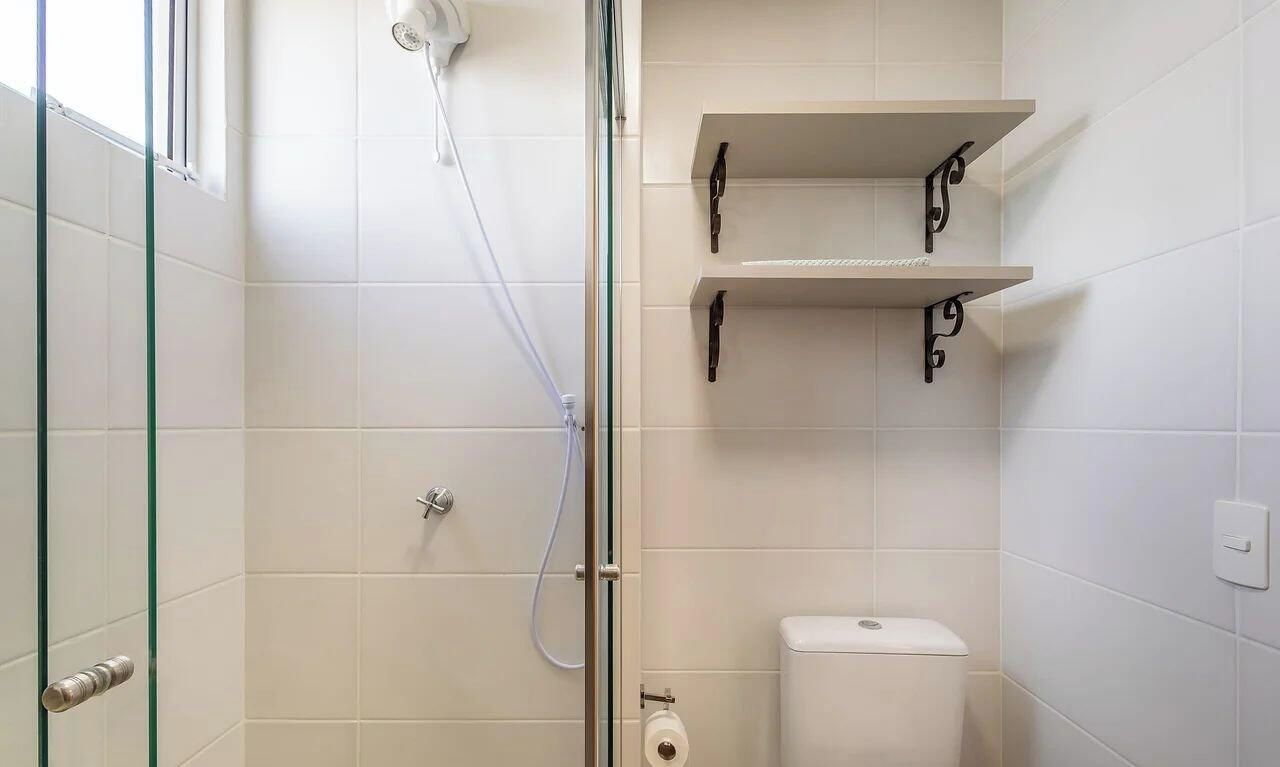 Un baño con ducha envuelta en vidrio, paredes de azulejos blancos, un inodoro y un estante con una toalla.