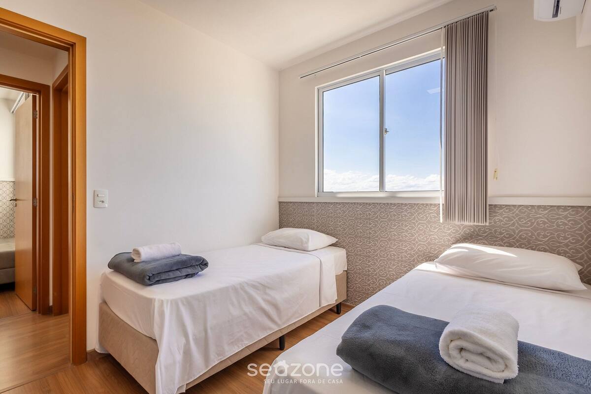 Quarto com duas camas, uma janela com cortinas e uma porta que dá para outro cômodo.