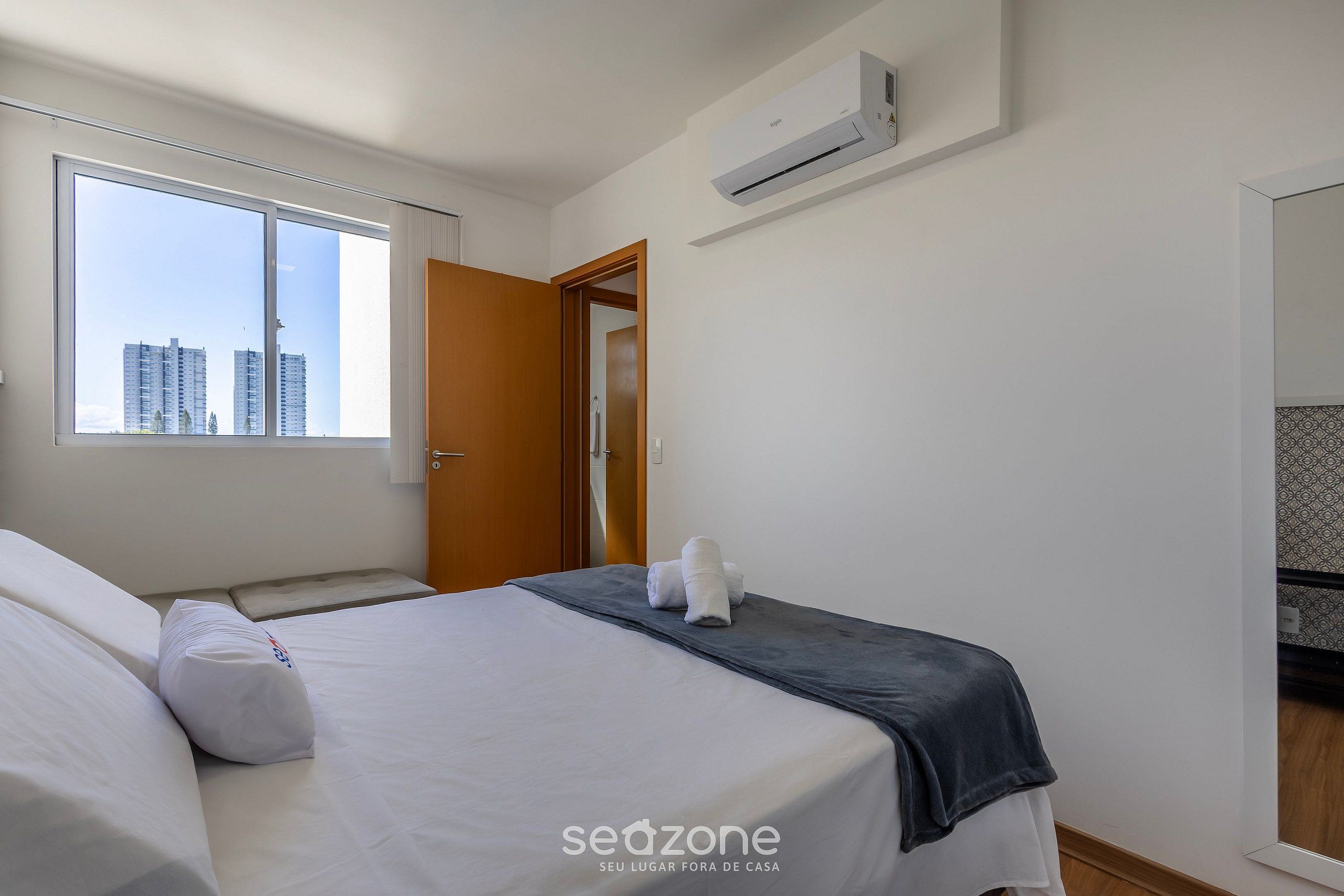 Habitación de hotel con un ventanal grande, una cama con ropa blanca, un edredón gris y una toalla sobre la cama.