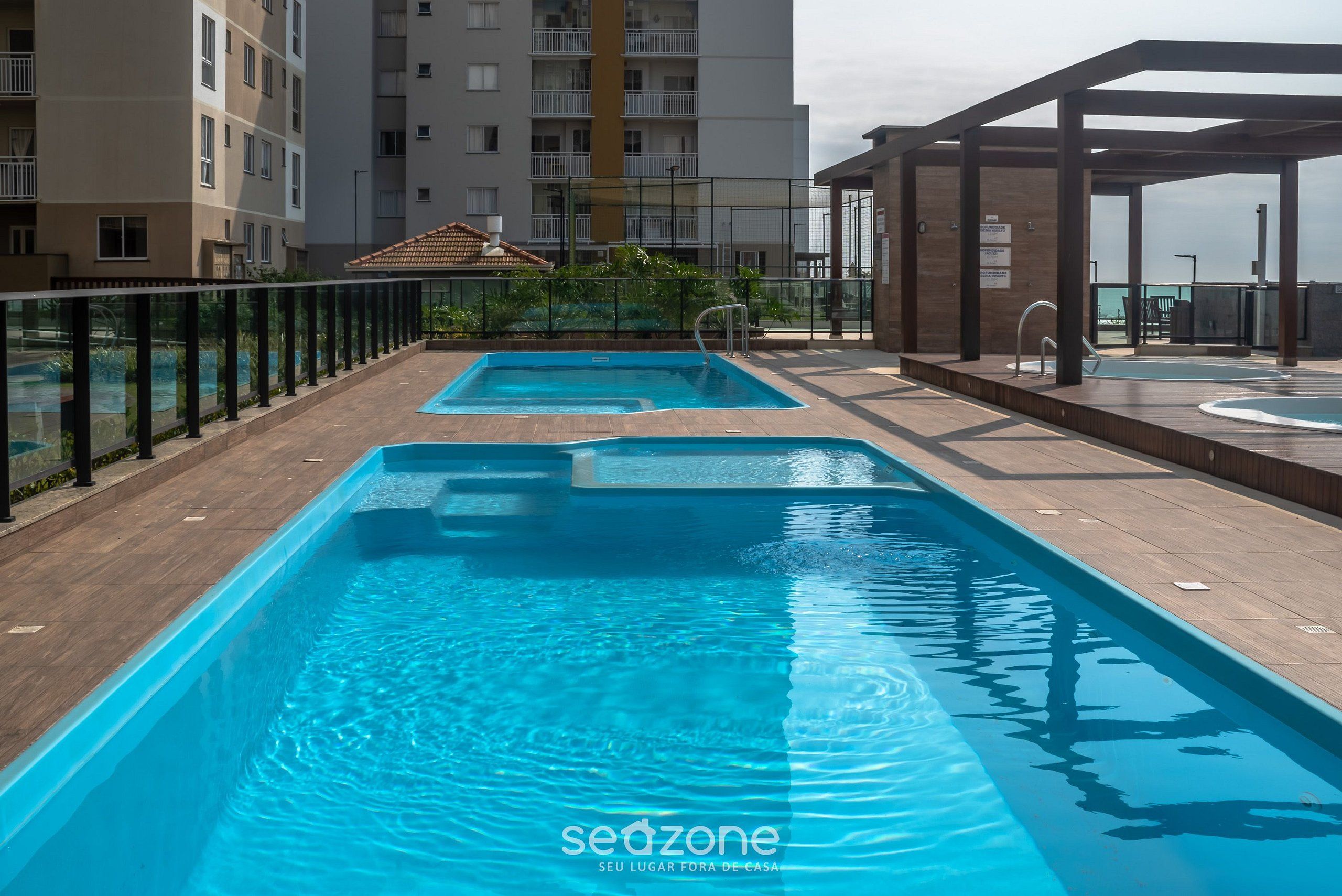 Una piscina en la azotea con un deck de madera, rodeada por una barandilla de vidrio y un edificio al fondo.