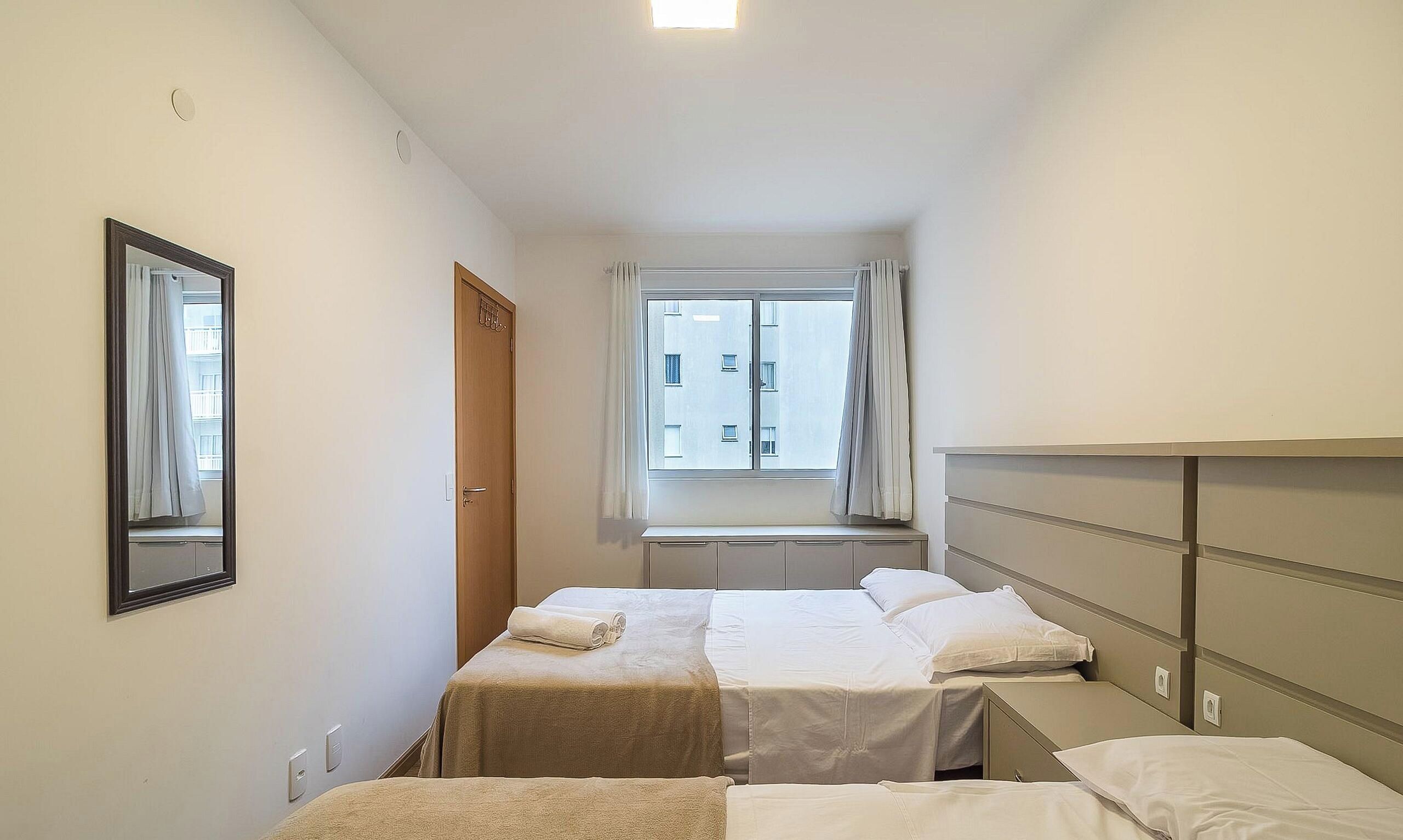 A hotel room with two beds, a window with curtains, a mirror, and a view of buildings outside.