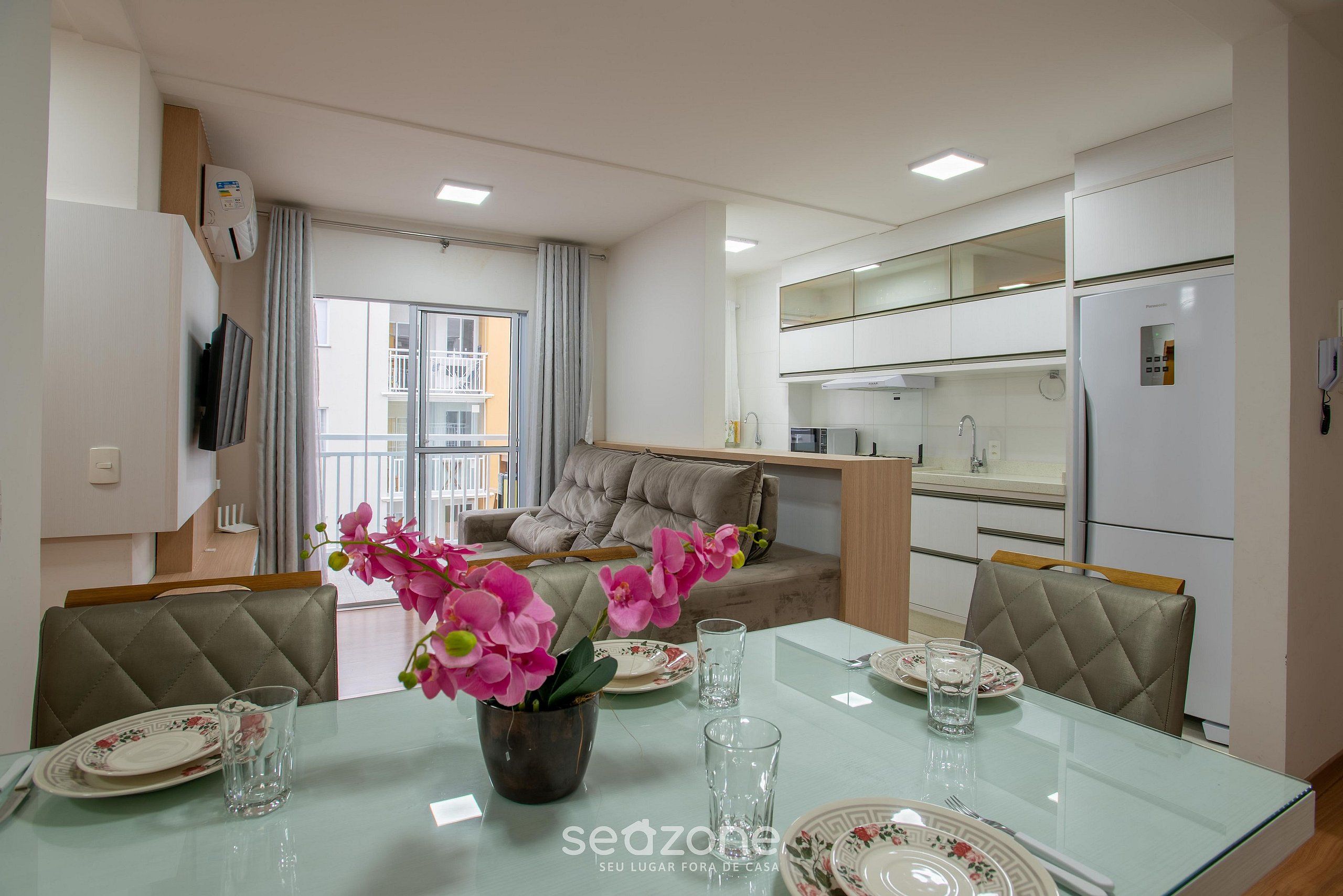 Una sala de estar moderna con una mesa de comedor de vidrio, un sofá y una cocina con armarios blancos.