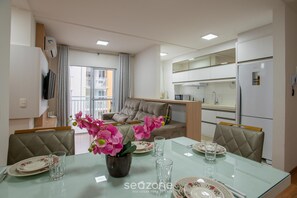 A modern living room with a glass dining table, a sofa, and a kitchen area with white cabinets. - VAC - Beachfront Condo in Itajuba Beach (Barra Velha)