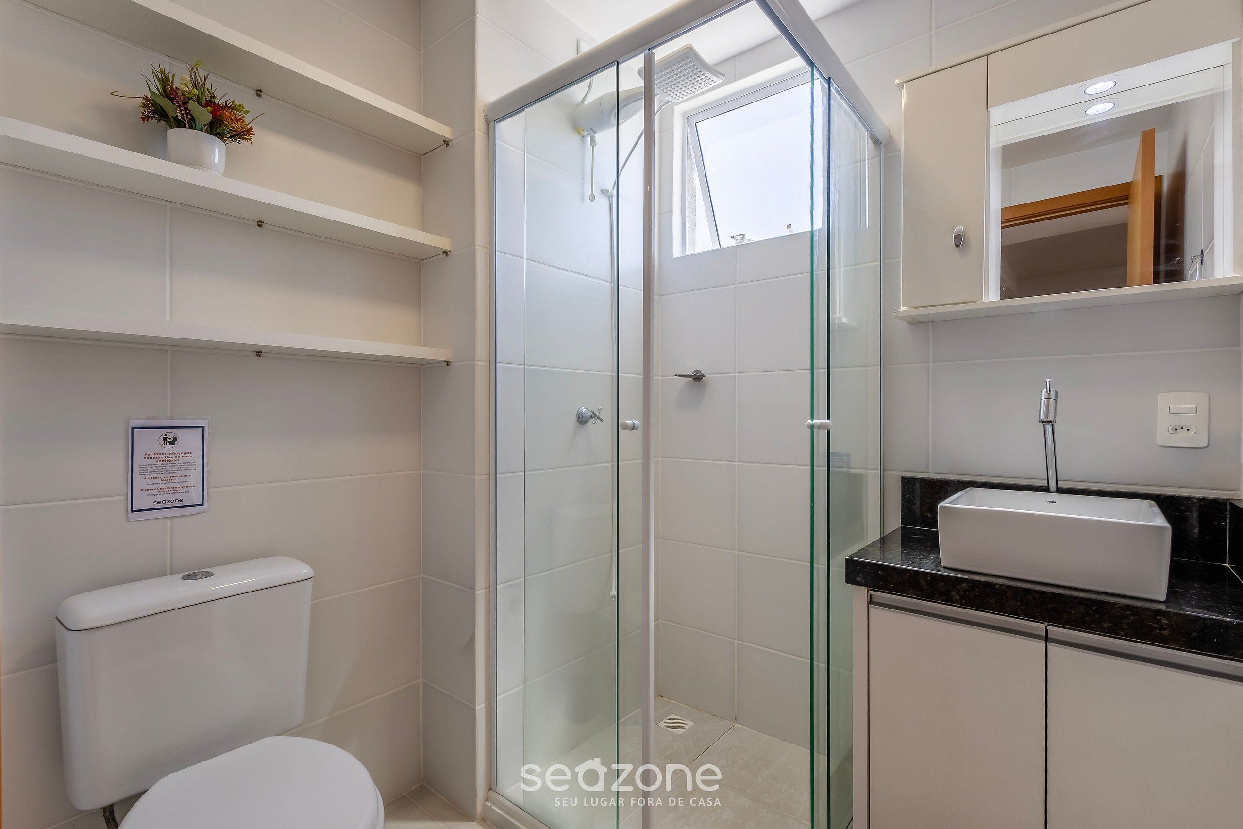 Un baño con cabina de ducha de vidrio, inodoro, lavamanos con espejo y estante con flores.