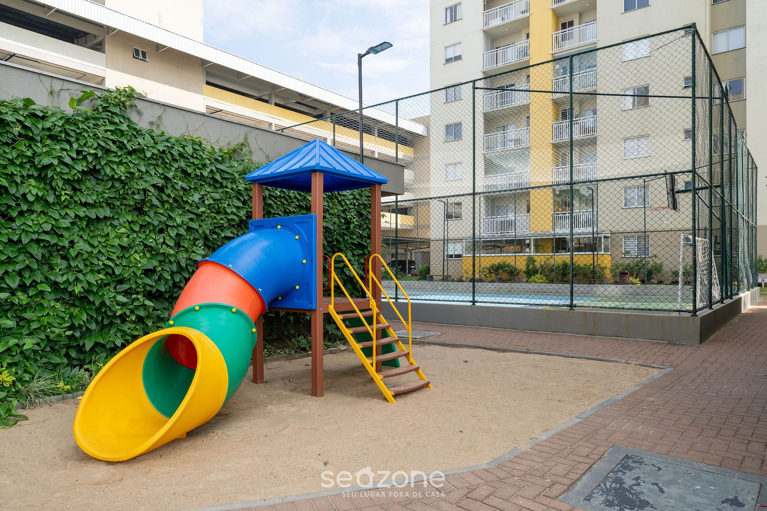 Un parque infantil con tobogán y estructura para escalar, rodeado por una cerca y un edificio al fondo.