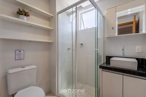 Un baño con cabina de ducha de vidrio, inodoro, lavamanos con espejo y estante con flores.
