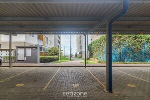 Uma área de estacionamento coberta com piso pavimentado, vigas de metal e vista para um prédio residencial ao fundo.