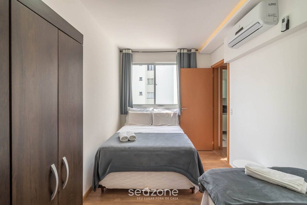 Quarto com cama, guarda-roupa, janela com cortinas e ar condicionado.