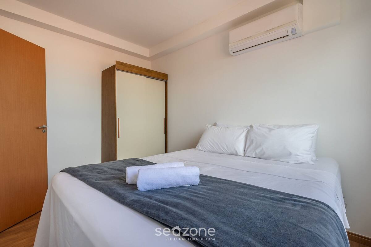 Quarto de hotel com uma cama, roupa de cama branca, um cobertor cinza, uma porta de madeira e um ar condicionado.