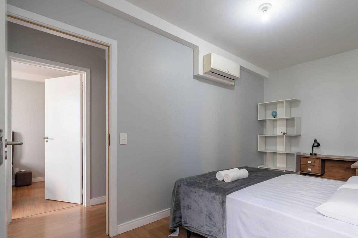 Quarto com cama, mesa de cabeceira, cômoda, estante e ar condicionado.