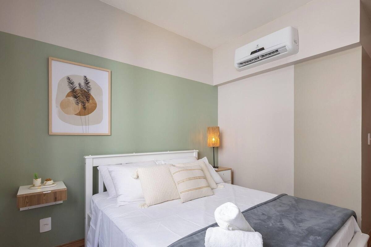 Quarto com cama, mesa de cabeceira, ar condicionado fixado na parede e uma obra de arte emoldurada.