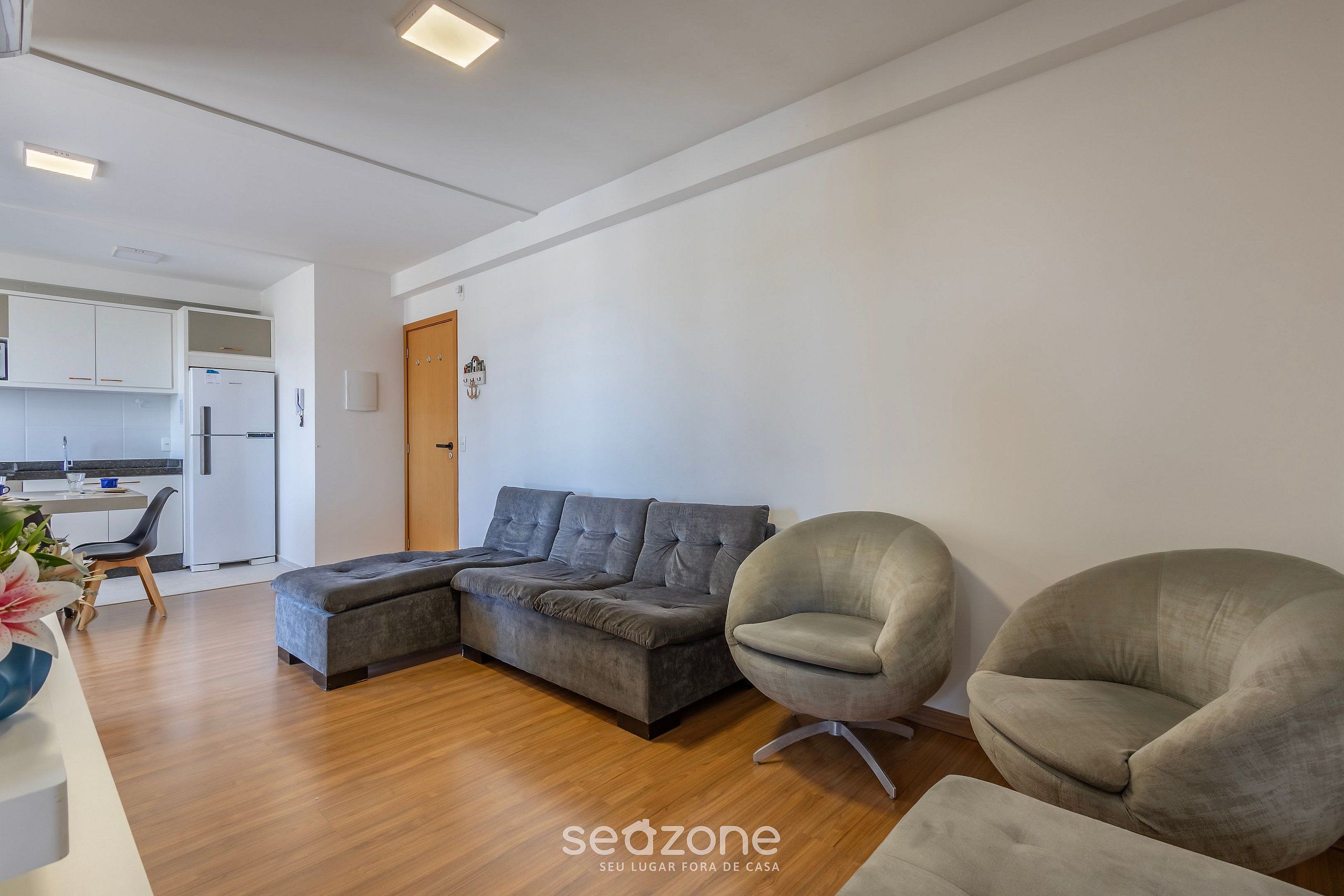 Un salon moderne avec un canapé d’angle, deux fauteuils, un plancher en bois et une cuisine en arrière-plan.