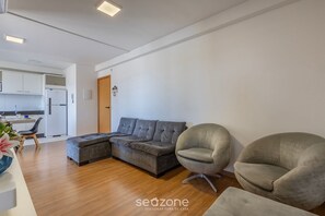 A modern living room with a sectional sofa, two armchairs, a wooden floor, and a kitchen in the background. - VAC - Beachfront Condo in Itajuba Beach (Barra Velha)