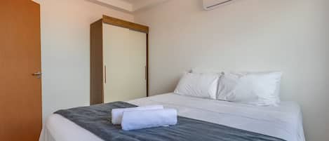 A hotel room with a bed, white linens, a grey blanket, a wooden door, and an air conditioning unit.