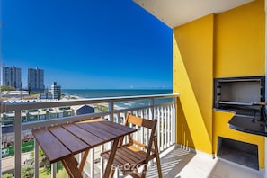 Un balcón con mesa y sillas, una parrilla y vistas al mar y edificios.