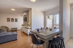 Una sala moderna con un sofá, un comedor con taburetes, una cocina con una isla grande y un televisor montado en la pared.