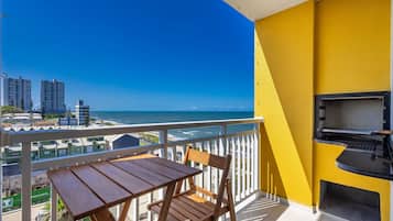 A balcony with a table and chairs, a barbecue grill, and a view of the sea and buildings.