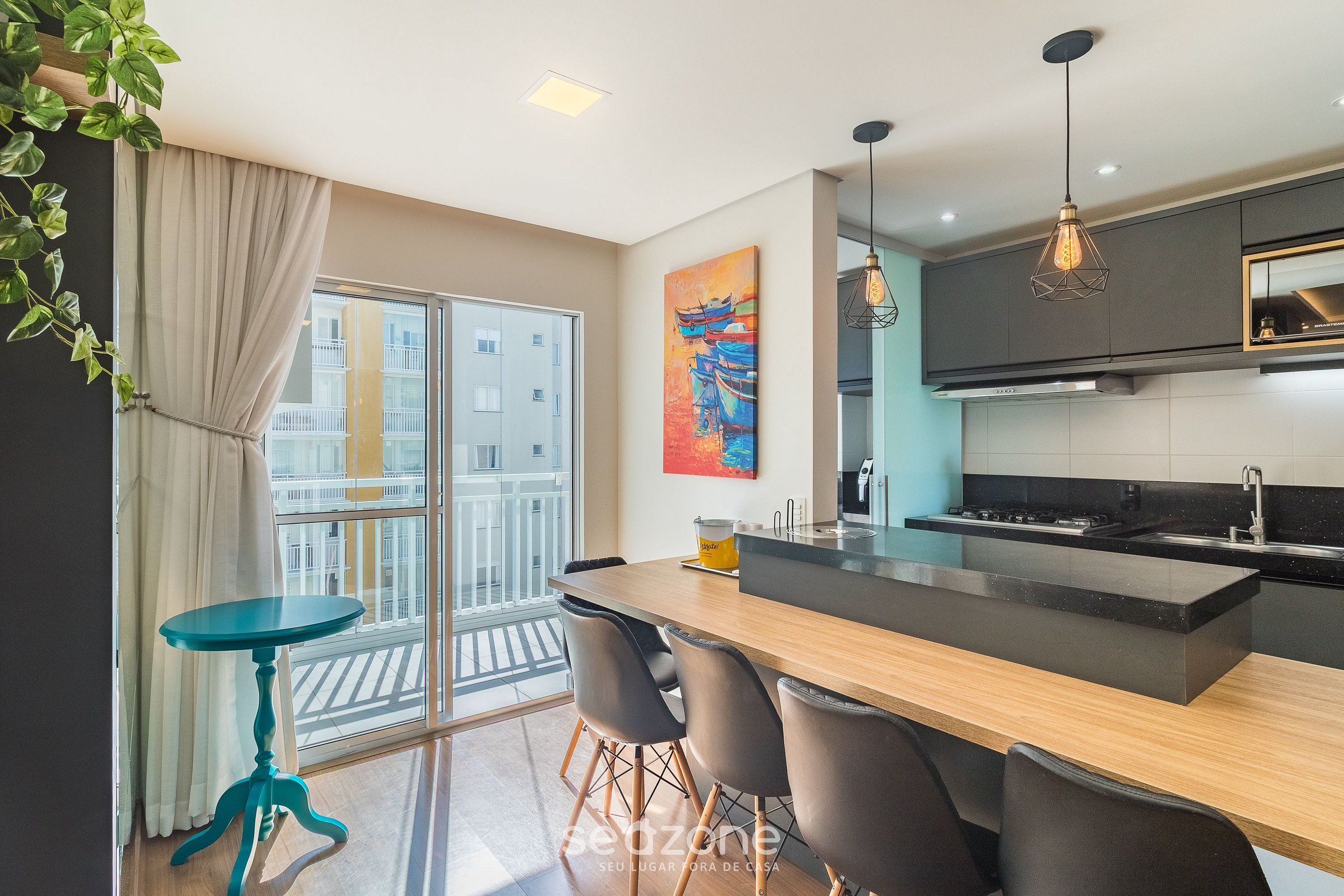 Uma cozinha moderna com uma ilha central, área de refeições com cadeiras, mesa azul e uma janela grande com cortinas.