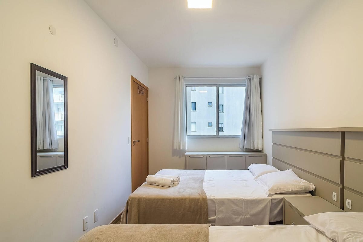 Quarto de hotel pequeno com duas camas, uma janela com cortinas, um espelho e um radiador.