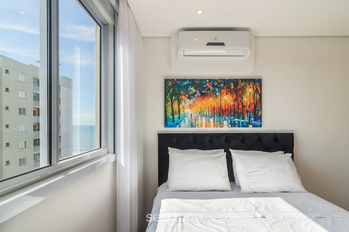 Quarto de hotel com uma janela grande, cama com roupa de cama branca, cabeceira preta e um quadro colorido na parede.