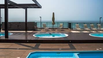 A rooftop pool area with lounge chairs and a hot tub, overlooking the sea.