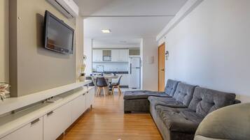 A modern living room with a grey sectional sofa, a flat-screen TV mounted on the wall, and a kitchen area in the background.