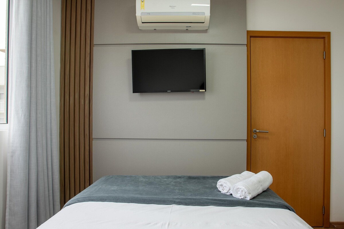 Una habitación de hotel con una cama, un televisor de pantalla plana montado en la pared, un equipo de aire acondicionado sobre el televisor y una puerta de madera.
