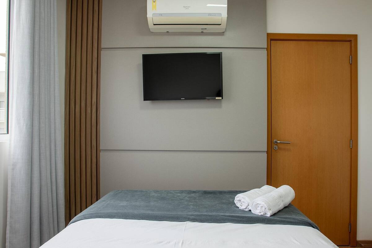 Quarto de hotel com cama, televisão de tela plana fixada na parede, ar-condicionado acima da TV e porta de madeira.
