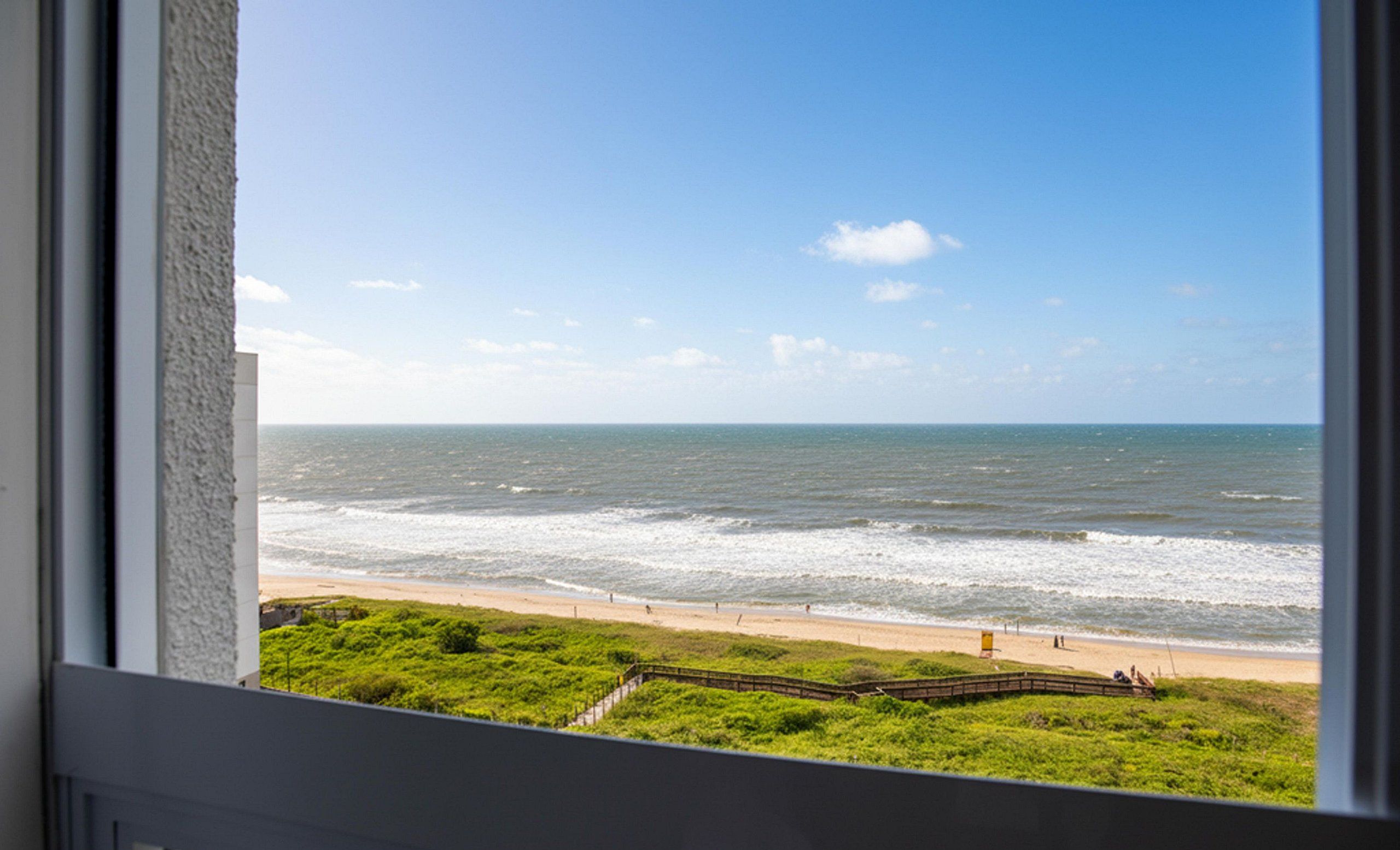 Vista del océano desde una ventana, con una playa y zona verde al frente.