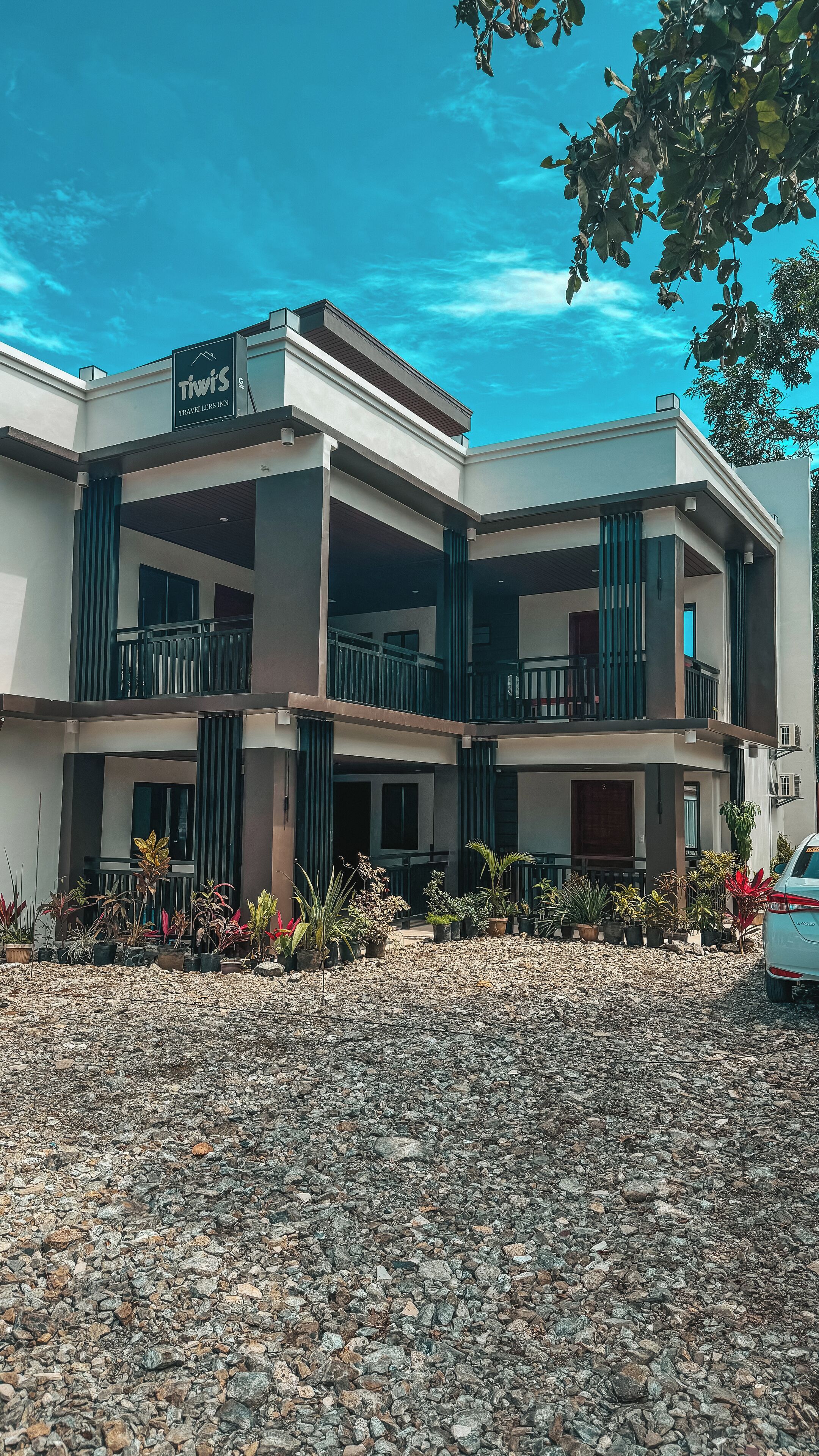 A modern building with a sign reading "Tivis's Travellers Inn". It features multiple balconies, a gravel driveway, and various potted plants.