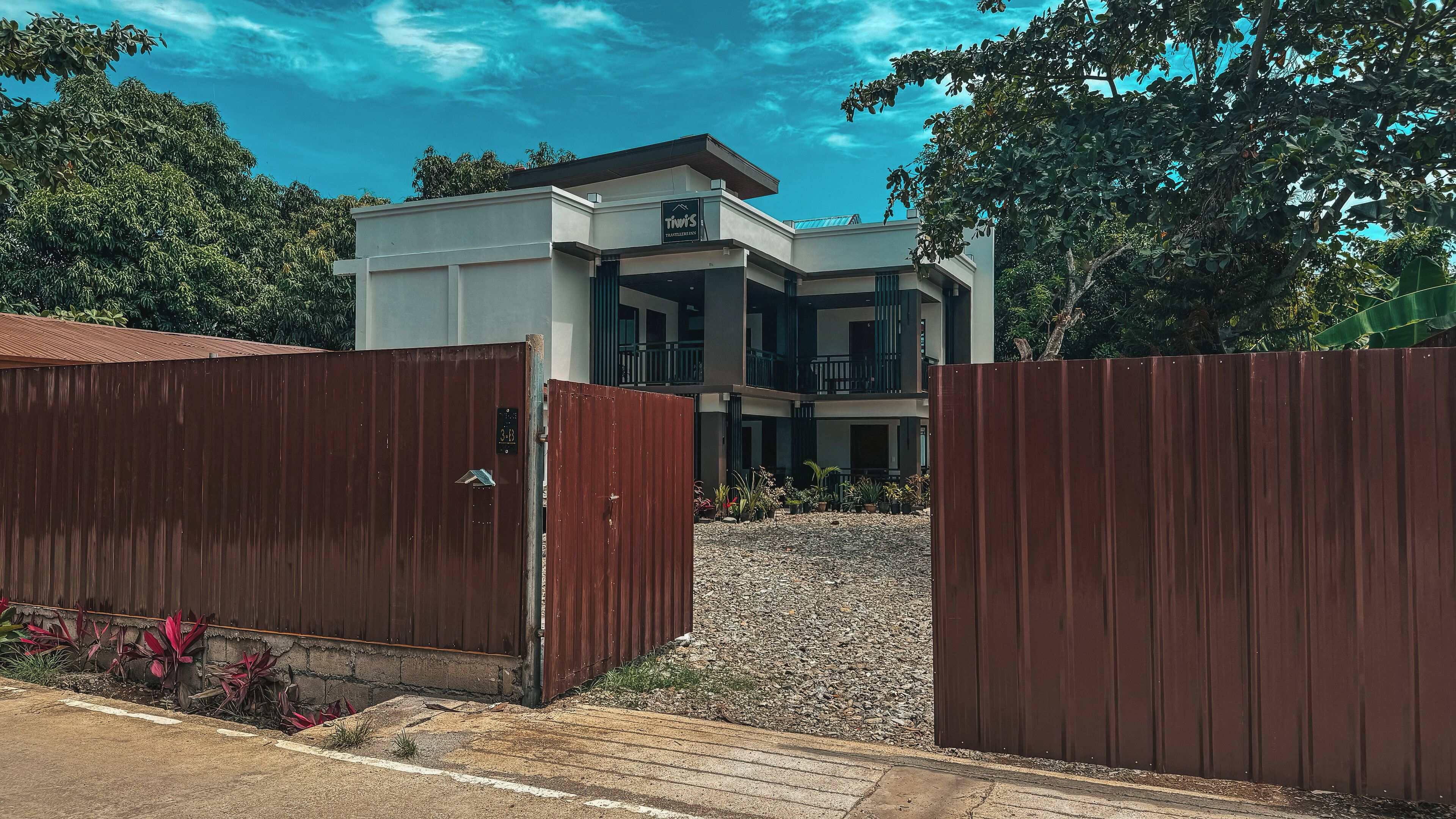 Um edifício moderno de dois andares com telhado plano, varanda no segundo andar e uma entrada gradeada com acesso por um caminho de cascalho.