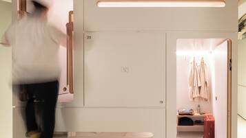 Quarto de hotel moderno com design minimalista, apresentando um amplo guarda-roupa com vários compartimentos, uma toucador embutido com espelho e uma pequena área de estar com uma mala vermelha.