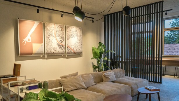 A modern living room with a beige sectional sofa, a small round table, and a bookshelf. There are large windows and a plant in the room. - soom Salzburg I Capsule Hotel (Salzburg)