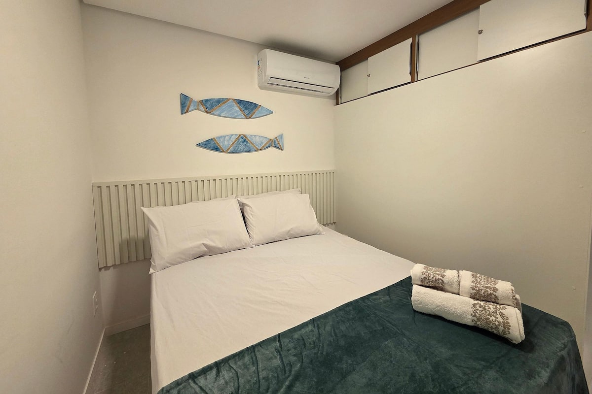 Ein Schlafzimmer mit einem Bett, weißer Bettwäsche, einem grünen Überwurf, zwei Wand-Klimaanlagen und zwei Wandbildern in Fischform.