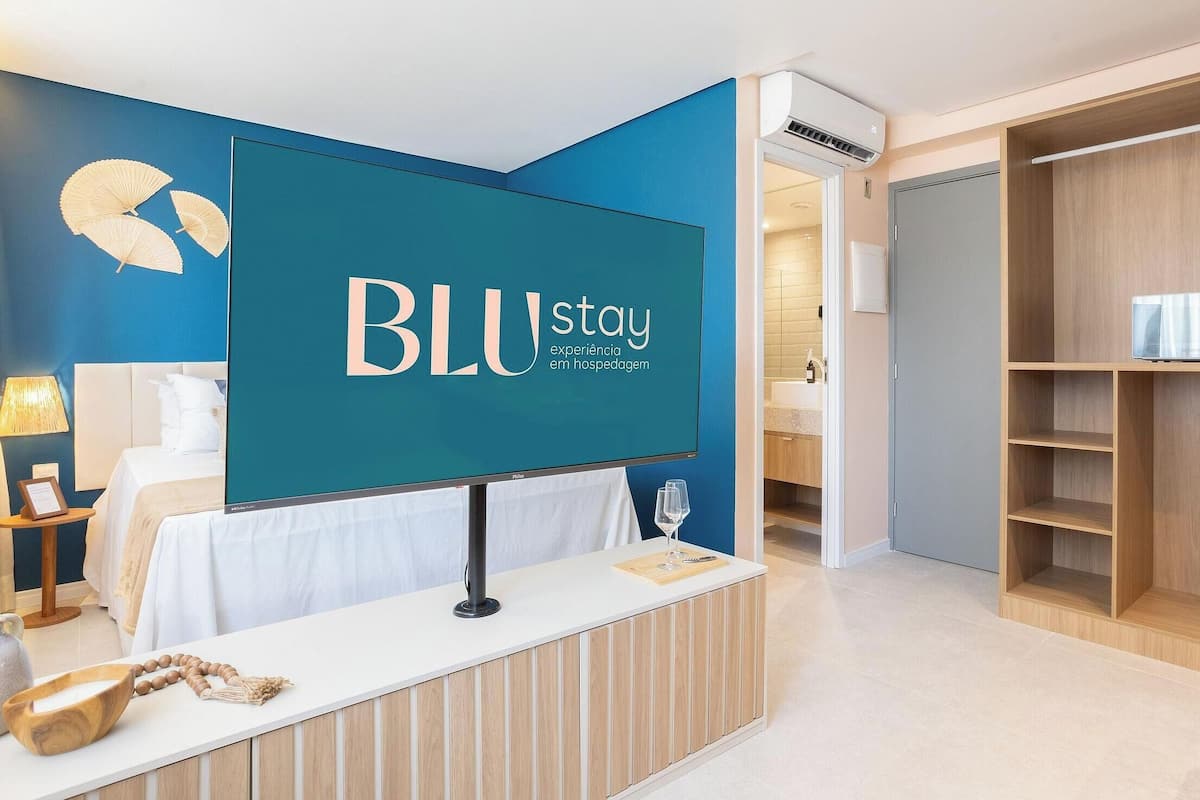 Ein Hotel-Empfangsbereich mit einem großen Schild, auf dem "BLU stay" steht. An der Rezeption stehen eine Lampe, ein Glas und eine Holbschüssel bereit. Im Hintergrund ist ein Bett mit weißer Bettwäsche zu sehen.