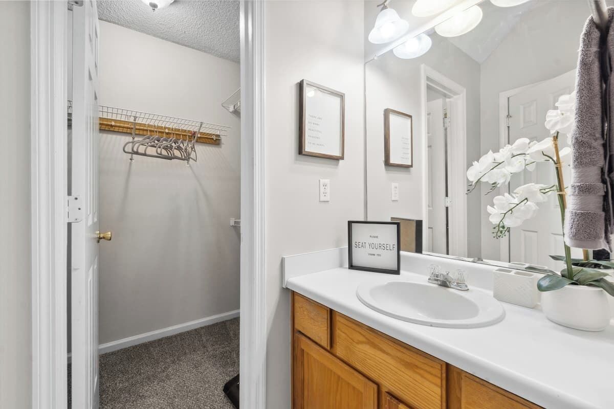 A modern bathroom with a white sink, wooden cabinets, and a mirror with lights. There are framed pictures on the wall and a sign that says "Please seat yourself."