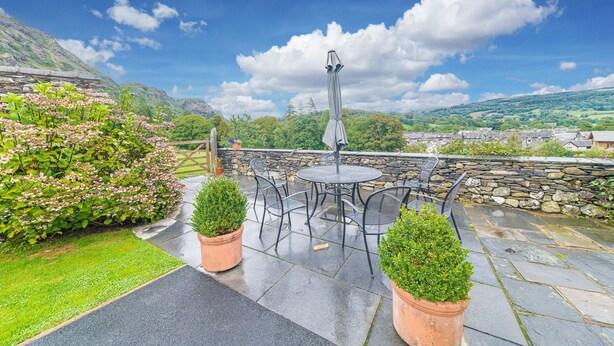 An outdoor patio with stone flooring, a round table, and chairs, surrounded by potted plants and a view of a village and hills.