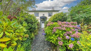 A house with a gravel pathway leading to the entrance, surrounded by lush greenery and flowering plants.