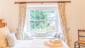A bedroom with a large window, a bed with white bedding, a bench with pillows, and a chair.