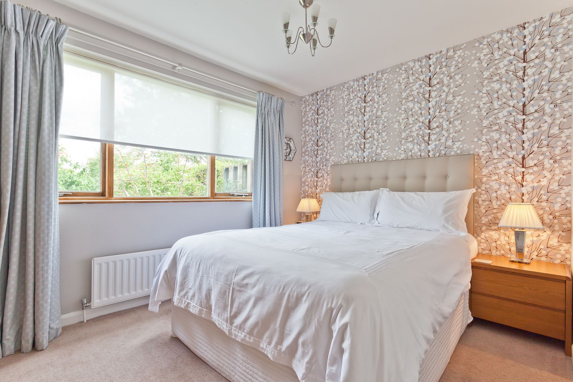 Une chambre avec un grand lit, un radiateur, une fenêtre avec des rideaux, un mur décoré d’un motif d’arbre et une table de chevet avec une lampe.