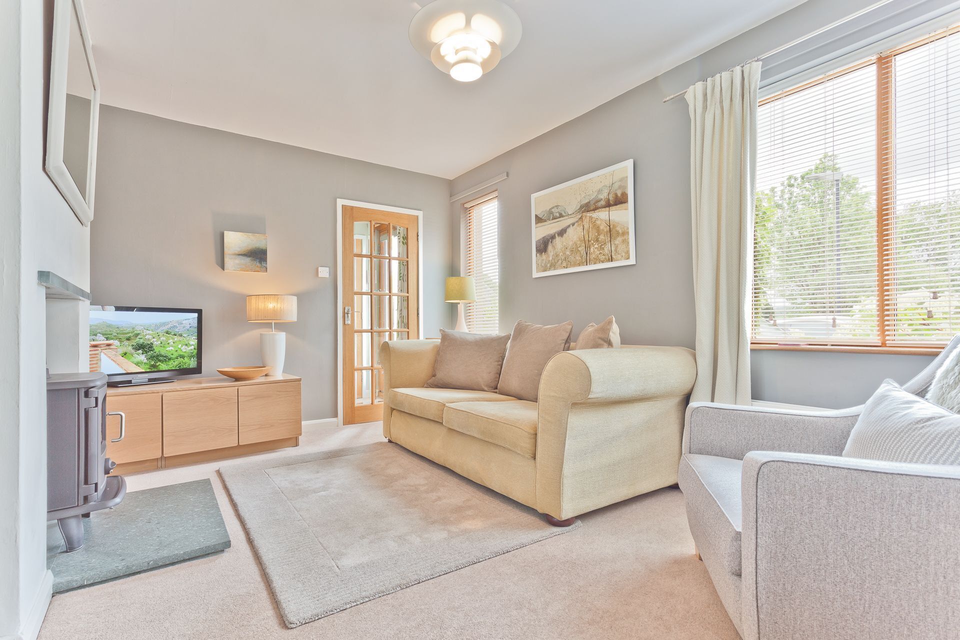 Een woonkamer met een beige bank, een grijze fauteuil, een televisie op een houten kast en een raam met jaloezieën.