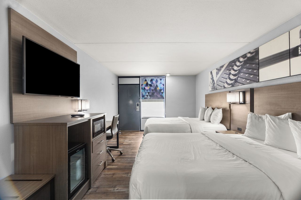 A hotel room with a flat-screen TV, a desk with a lamp, two beds, a wardrobe, and a large window.